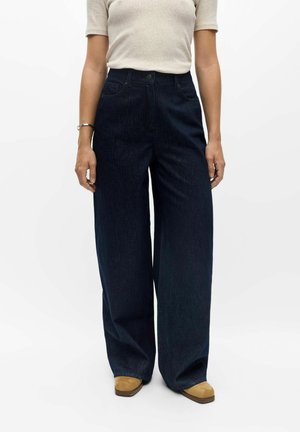 Mulher vestindo jeans escuros de perna larga, uma blusa leve de mangas curtas, sapatos cor de creme e uma pulseira, em pé contra um fundo branco liso.