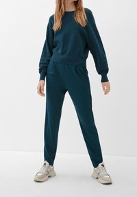 Kobieta w turkusowym, strukturowanym swetrze z długim rękawem i dopasowanych spodniach, stojąca na tle czystej białej ściany, w białych, masywnych trampkach.