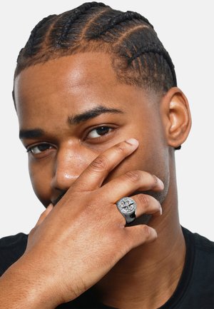 Silver and black ring with a circular design, featuring intricate patterns and a textured finish, worn on a hand against a plain background.