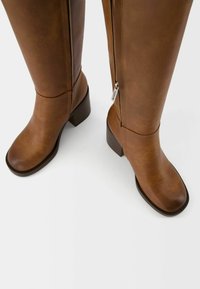 Bottes marron montant jusqu'au genou en cuir lisse, dotées d'une fermeture éclair latérale et d'un talon bloc. Le design est simple, sans motifs ni accents supplémentaires.