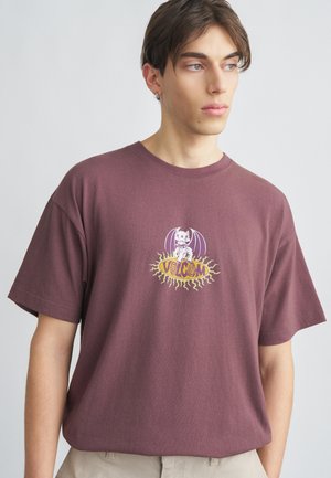 Mladý muž nosiaci fialovoružové tričko s grafickým motívom komiksového psa s netopierími krídlami a nápisom „Volcom“, pozerajúci sa bokom na jednoduchom pozadí.