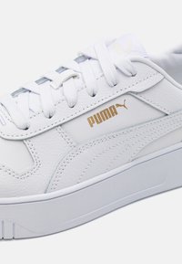 Fehér bőr sneaker texturált felsőrésszel, lapos fehér fűzőkkel, arany "PUMA" logóval az oldalon, és tömzsi fehér talppal.