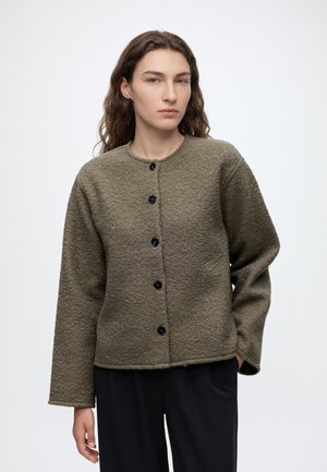 Jong persoon met lang golvend haar, gekleed in een getextureerd olijfgroen jasje met knopen en een zwarte broek tegen een effen achtergrond.