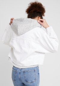 Chaqueta vaquera blanca recortada con detalles desgastados, que lleva una sudadera con capucha gris debajo. Completan el conjunto unos vaqueros azules de talle alto.