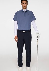 Heren polo met korte mouwen in lichtblauw met logo; daaronder een wit shirt met lange mouwen, gecombineerd met donkerblauwe broek en witte schoenen, terwijl hij een golfclub vasthoudt.
