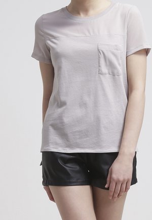 T-shirt grigia chiara con maniche corte e taschino sul petto, caratterizzata da una morbida texture e un pannello superiore leggermente trasparente, abbinata a pantaloncini neri.