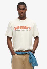 T-shirt color crème avec le logo "SUPERDRY" en orange. Tissu en coton doux, manches courtes et coupe décontractée. Ceinture marron avec une chemise en denim nouée à la taille.