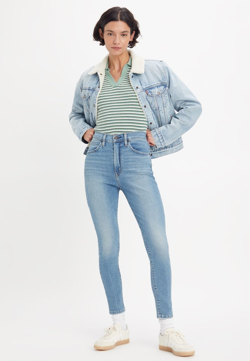 Frau steht mit Händen in der Hüfte, trägt eine hellblaue Jeansjacke mit weißem Fleecekragen, gestreiftes grünes Shirt, hochgeschnittene Jeans und weiße Sneaker.