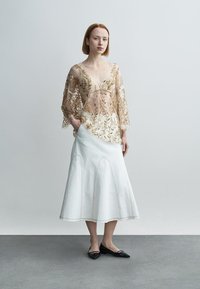 Διαφανές μπεζ τοπ με χρυσή κεντητική με λουλούδια, συνδυασμένο με λευκή φούστα με πιέτες και φουσκωτή γραμμή, συνοδευόμενο από μαύρα φλατ παπούτσια.