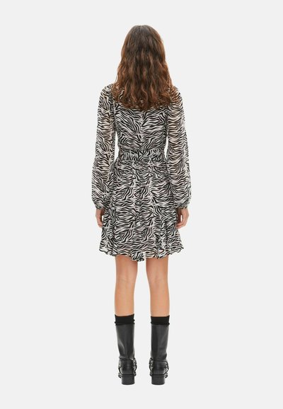 Mujer con cabello ondulado vestida con un vestido de manga larga con estampado de cebra en blanco y negro y botas negras hasta media pantorrilla, de pie mirando hacia atrás.