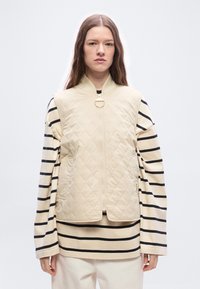 Γυναίκα με μακριά καστανά μαλλιά που φοράει μπεζ γιλέκο με καπιτονέ ύφασμα πάνω από μπεζ και μαύρο ριγωτό μακρυμάνικο πουκάμισο και ανοιχτόχρωμο παντελόνι.