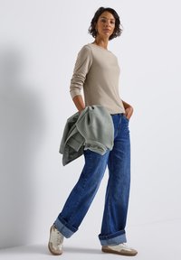 Beige langemouwen top, blauwe straight-leg jeans en creamkleurige sneakers. Een groene sweatshirt hangt over de arm. Eenvoudig ontwerp, relaxte pasvorm.