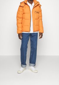 Pomarańczowa kurtka puchowa z zamkiem błyskawicznym i zatrzaskami, przednią kieszenią oraz kapturem. Nosi się ją na białą koszulę, w zestawie z podwiniętymi niebieskimi dżinsami i białymi tenisówkami.