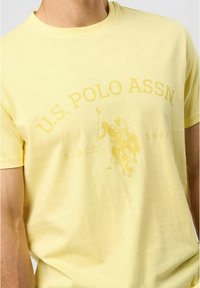 Tricou din bumbac galben cu o textură moale, decolteu rotund și un grafic subtil cu jucători de polo, având textul „U.S. POLO ASSN.”.