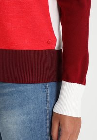 Suéter rojo y blanco con textura de punto, que presenta un diseño de bloques de color, puños acanalados y un pequeño detalle de botón de marca en el lado.