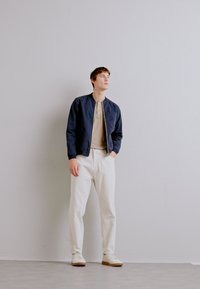 Chaqueta de bombardero azul marino, camisa henley beige, pantalones blancos texturizados y zapatillas blancas. El modelo está de pie contra un fondo gris claro.