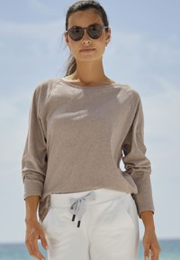 Svijetlosmeđi dugi rukav top s raglan rukavima, u kombinaciji s bijelim hlačama na vezanje. Model nosi tamne sunčane naočale i stoji na otvorenom.