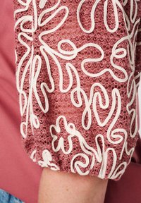 Roze blouse met een textuur, geborduurde mouw met witte draaiende patronen. De stof is zacht met een geplooide manchette.