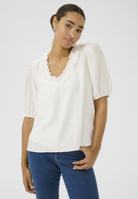 Haut blanc à manches courtes avec un décolleté en V à volant, fabriqué dans un tissu léger, offrant une coupe décontractée et une texture douce. Porté avec un jean bleu.