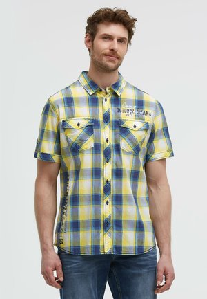 Mann trägt ein gelb-blau kariertes Kurzarmhemd mit Brusttaschen und blaue Jeans, stehend vor einem einfarbigen hellen Hintergrund.