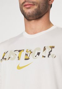 Biała bawełniana koszulka z krótkim rękawem z logo "Just Do It" w różnych wzorach kamuflażu oraz żółtym symbolem Nike. Okrągły dekolt.