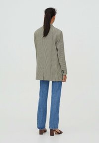 Blazer à motif pied-de-poule en beige clair et noir, coupe ajustée avec une fente unique, porté avec un jean bleu évasé et des sandales à talons marron.