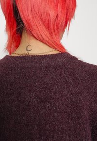 Mörklila ulltröja med rund halsringning, med en texturerad stickning. En guldkedja och en liten halvmåne-tatuering syns.