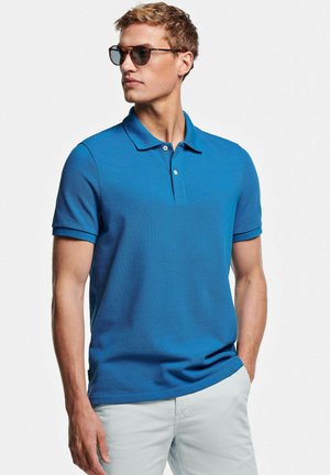 Jonge man met schildpadkleurige zonnebril, een blauw poloshirt met korte mouwen en lichtgekleurde broek, poserend met één hand in zijn zak.