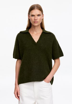 Femme aux cheveux blonds raides portant un pull vert foncé à manches courtes avec col et un pantalon blanc, debout avec une main dans la poche.