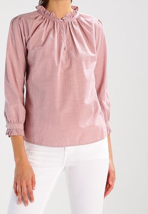 Femme portant un chemisier rose clair à manches longues avec col et poignets volantés, associé à un pantalon blanc, debout devant un fond uni.