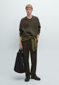 Brauner gerippter Pullover, olivgrüner Wickelmantel, dunkle Hose, schwarze Wildleder-Tasche mit hellen Trägern, schwarze Schuhe, schlicher Hintergrund, lässiger Look.