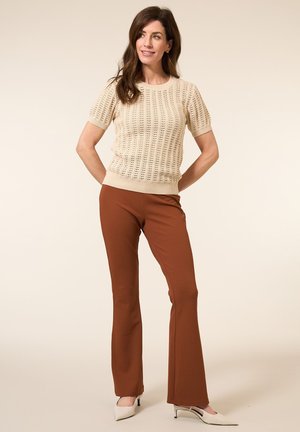 Gebreide beige top met korte mouwen, open patronen en een geribbelde zoom, gecombineerd met roestkleurige wijde broek; model draagt witte hakken.