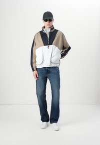 Fargeblokket vindjakke i beige, marineblå og hvit. Har høy krage, front lommer og en lett, glattere tekstur. Båret med blå jeans og hvite sneakers.
