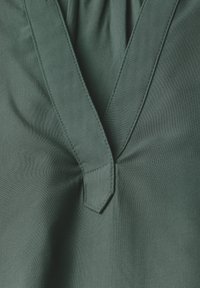 Tessuto verde con una texture liscia, caratterizzato da un design a V, dettagli arricciati e accenti cuciti lungo il colletto.