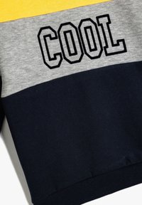 Sudadera con bloques de color en amarillo, gris claro y azul marino, que presenta la palabra "COOL" en letras grandes de contorno negro.