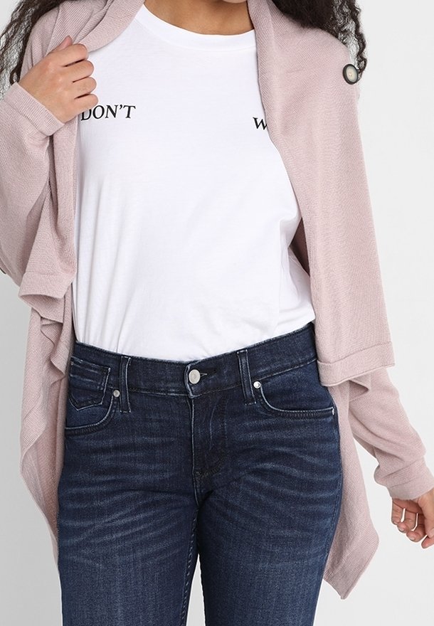 Hellrosa Strickjacke mit strukturierter Oberfläche, getragen über einem weißen T-Shirt mit Schriftzug, kombiniert mit dunkelblauen Jeans und einem Knopfdetail.