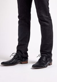 Zwarte leren sneakers met een golvende textuur, puntige neus en vetersluiting. Gecombineerd met donkere jeans die een slim fit laten zien.
