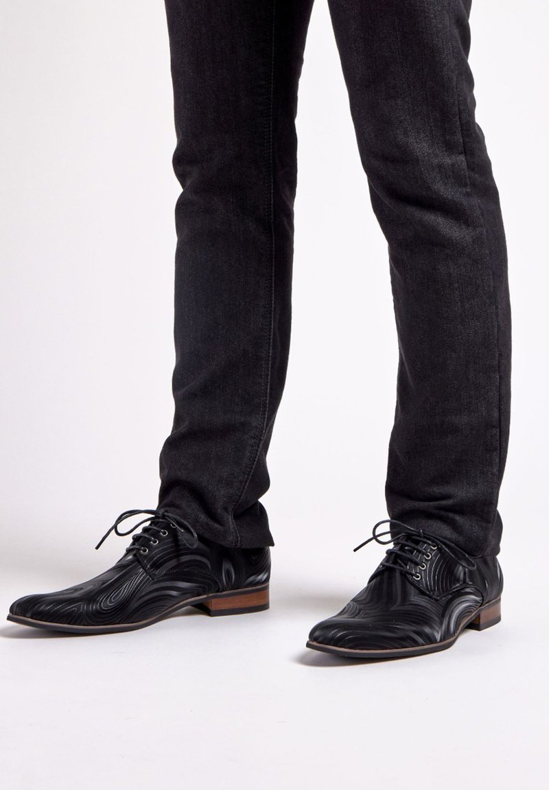Zwarte leren sneakers met een golvende textuur, puntige neus en vetersluiting. Gecombineerd met donkere jeans die een slim fit laten zien.