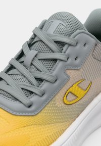 Grijze en gele sneaker met een mesh bovenkant, getextureerde gradiënten, platte veters en een geel logo tegen een gladde, grijze trim.