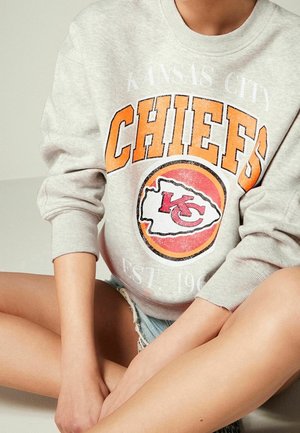 Personne portant un sweat-shirt gris Kansas City Chiefs et un short en jean, assise en tailleur avec les mains posées sur les genoux.