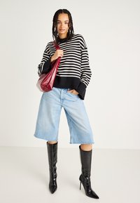 Sweater riscado em preto e branco, culotes de denim azul claro com bainha desfiada, e botas pretas de bico fino. Segurando uma mala de couro bordô.