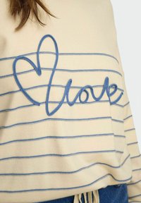 Pull beige à rayures horizontales bleues et fil bleu en relief formant le mot "love" avec un cœur au début, porté avec un jean bleu.