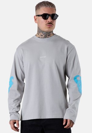 Chemise grise à manches longues avec un col rond, ornée d'un graphique bleu sur la manche gauche et d'un petit logo blanc sur la poitrine.