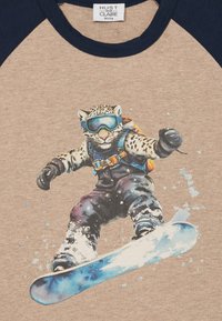 Bež majica s grafikom leoparda koji vozi snowboard, noseći naočale i crno skijaško odijelo s plavim akcentima te žutom ruksakom.
