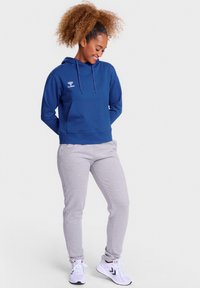 Marineblaue Hoodie mit Fronttasche, verstellbaren Kordeln und weißem Logo; kombiniert mit hellgrauen Jogginghosen und weißen Sneakers.