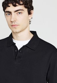 Polo nero con tessuto testurizzato, maniche corte e una chiusura a tre bottoni. Indossato sopra una maglietta bianca, con colletto classico.