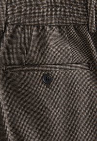 Gros plan sur un tissu à motif pied-de-poule marron et beige avec une poche à rabat boutonnée sur un vêtement sur mesure.