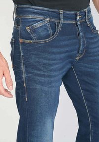 Jeans en denim bleu foncé avec une coupe slim. Les caractéristiques incluent des coutures visibles, cinq poches et une texture lisse.