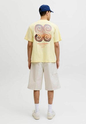Man in blue cap, yellow t-shirt with red and black swirl design and text, white knee-length shorts, white socks, and white sneakers, facing away.