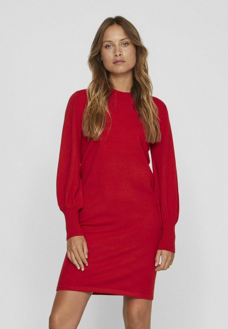 Donna con capelli lunghi che indossa un vestito a maglia rosso a maniche lunghe, in piedi contro uno sfondo grigio chiaro semplice.